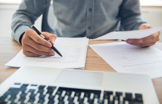Unrecognizable businessman sitting at table holding ballpoint pen and papers and checking documents in office
